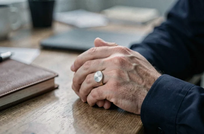 Mains masculines portant une chevalière argentée sur l'auriculaire et une alliance dorée, posées sur un bureau en bois avec carnet en cuir